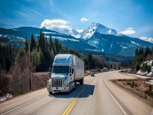 A moving truck driving across a country road, with mountains in the background.